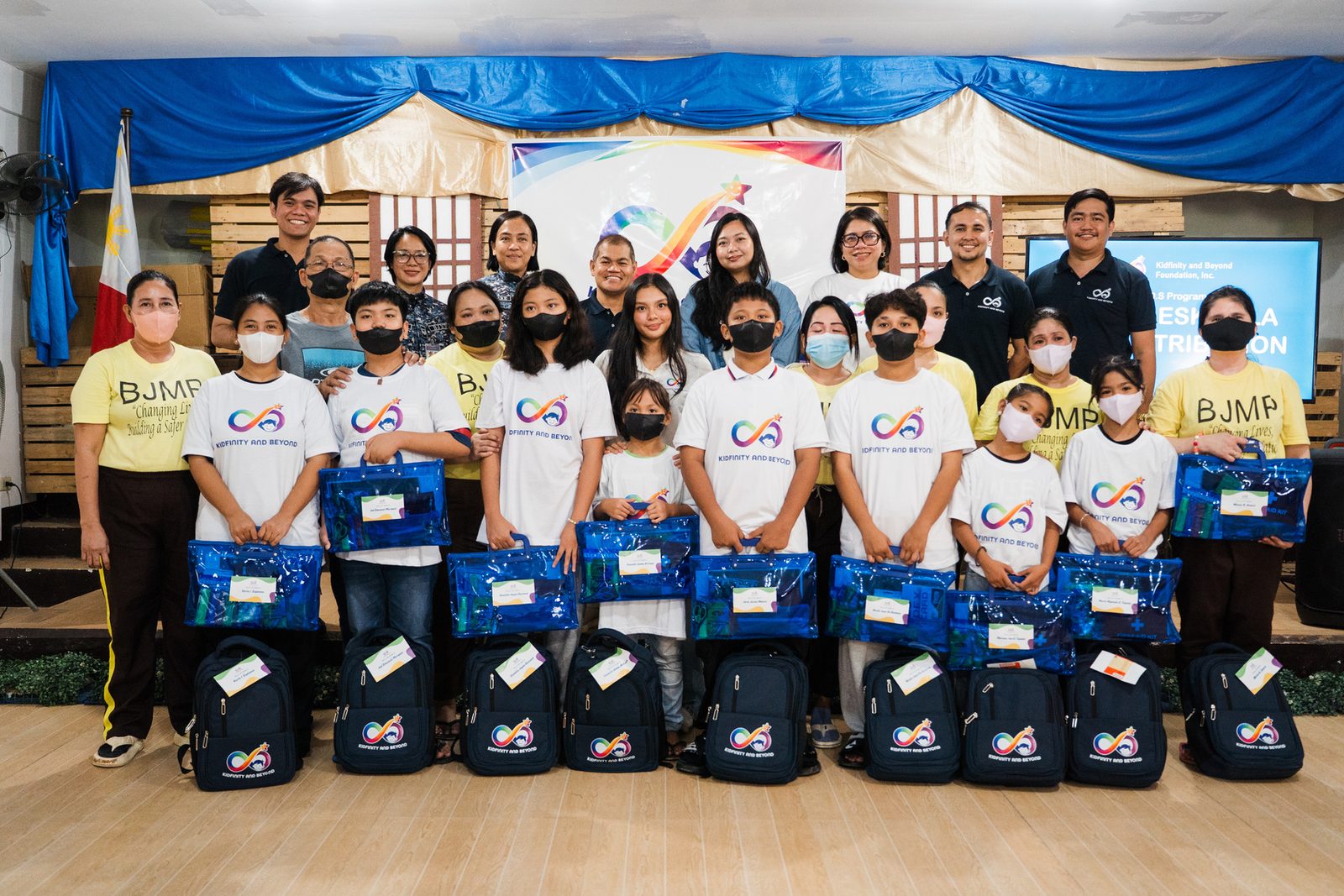 Child beneficiaries in Kidfinity shirts stand in rows holding blue document bags, with backpacks at their feet, alongside Foundation staff and jail personnel.