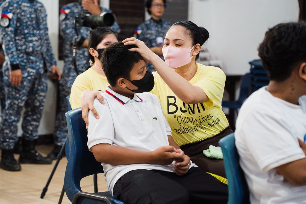 A mother in a yellow shirt gently touches her son’s head as he sits in a chair.